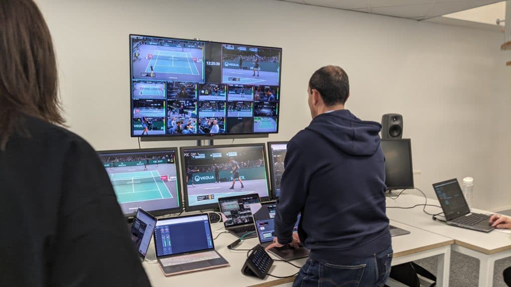 A photo of a director during a virtualized live production, with a multiviewer on the wall in a TV screen, laptops and monitors showing the production
